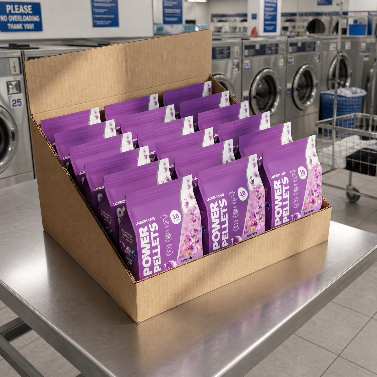 A cardboard display box filled with multiple purple bags of laundry detergent pellets, placed on a stainless steel table in a laundrette with washing machines in the background.